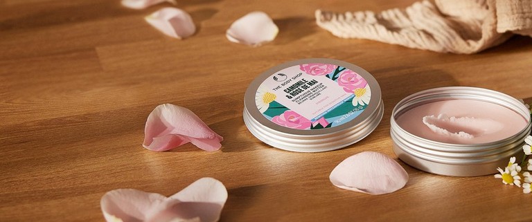 An open screw lid tin of rose de mai camomile makeup cleansing butter on a wooden table with rose petals and camomile flowers next to it.
