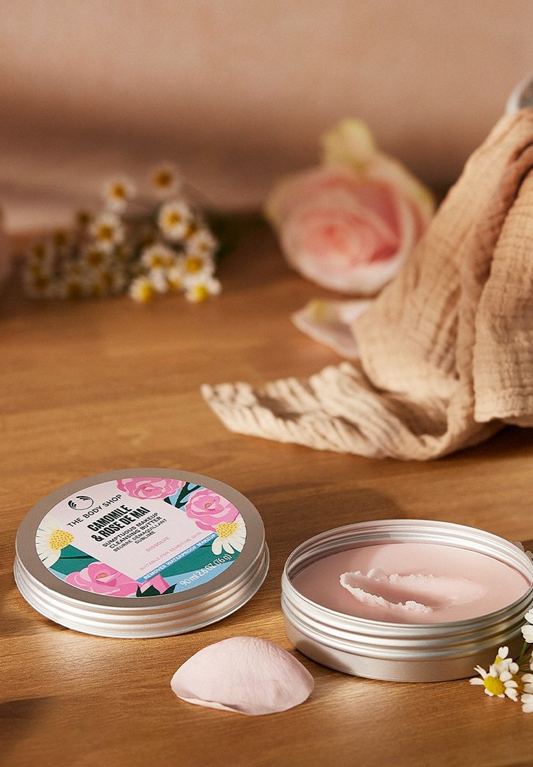 An open screw lid tin of rose de mai camomile makeup cleansing butter on a wooden table with rose petals and camomile flowers next to it.